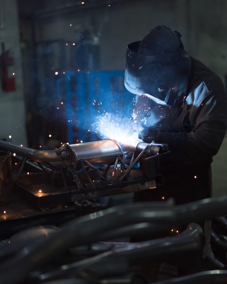 Pionowe ujęcie spawacza w ciemnym ubraniu i przyłbicy, pracującego w słabo oświetlonej hali przemysłowej. Spawa metalowe rury lub elementy wydechu. Łuk spawalniczy jest jaskrawo niebieski i otoczony iskrami.