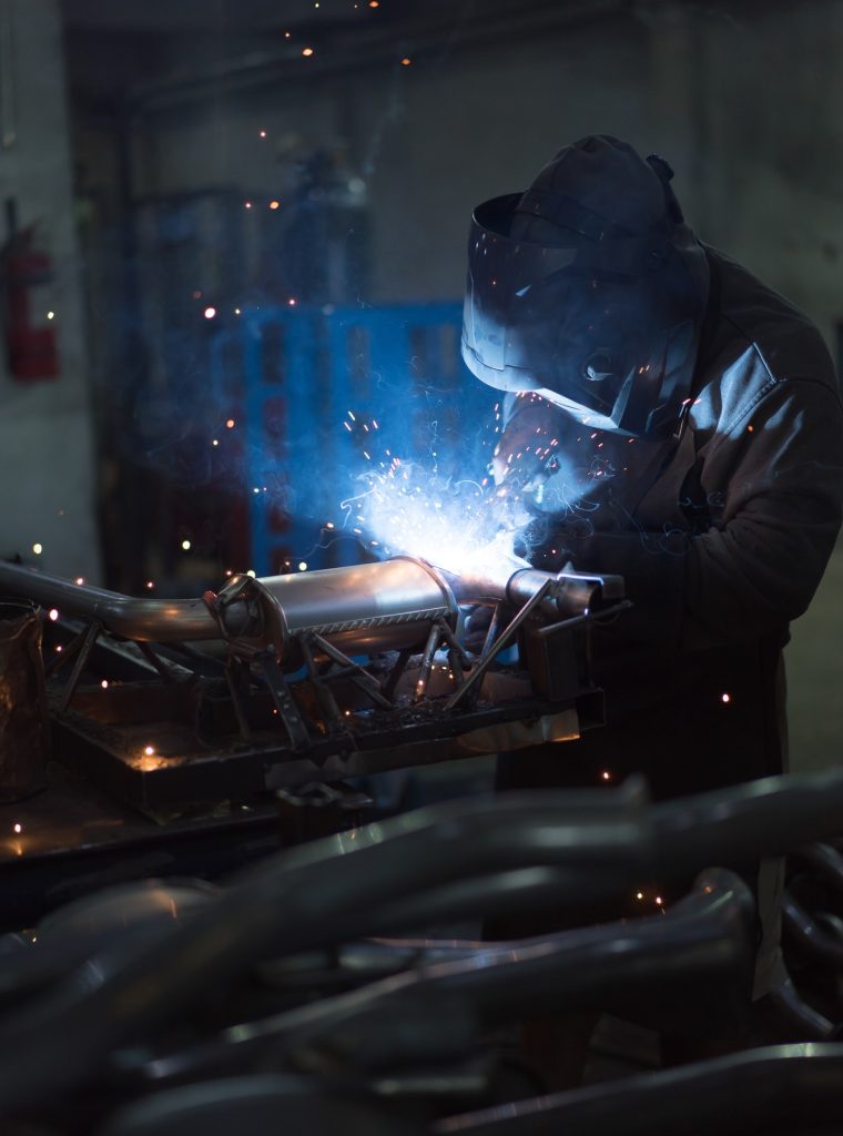 Pionowe ujęcie spawacza w ciemnym ubraniu i przyłbicy, pracującego w słabo oświetlonej hali przemysłowej. Spawa metalowe rury lub elementy wydechu. Łuk spawalniczy jest jaskrawo niebieski i otoczony iskrami.